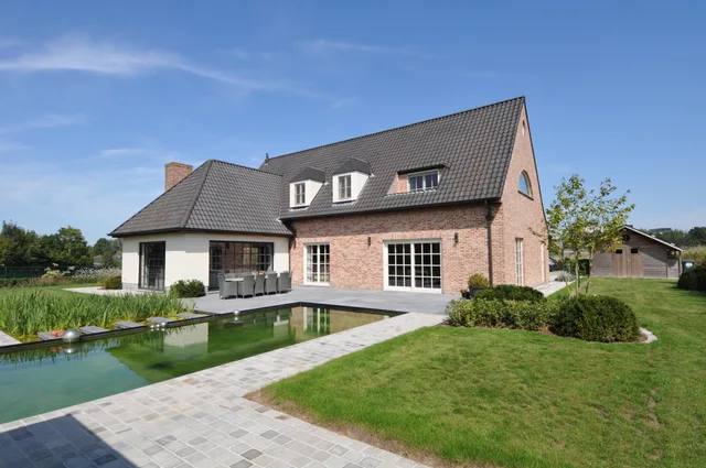 Moderne bakstenen villa met grote raampartijen, terrasoverkapping en aangrenzende vijver in groene omgeving
