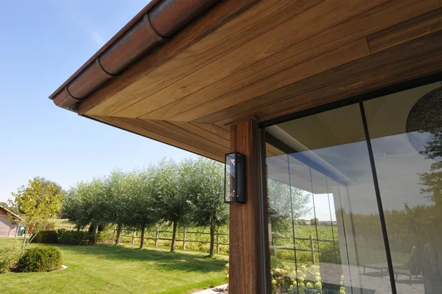 Houten terrasoverkapping met grote glazen schuifdeuren, uitzicht op aangelegde tuin met bomen