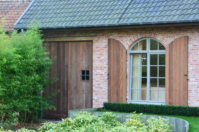 Bakstenen woning met houten deuren en raam met luiken, groene haag in tuin