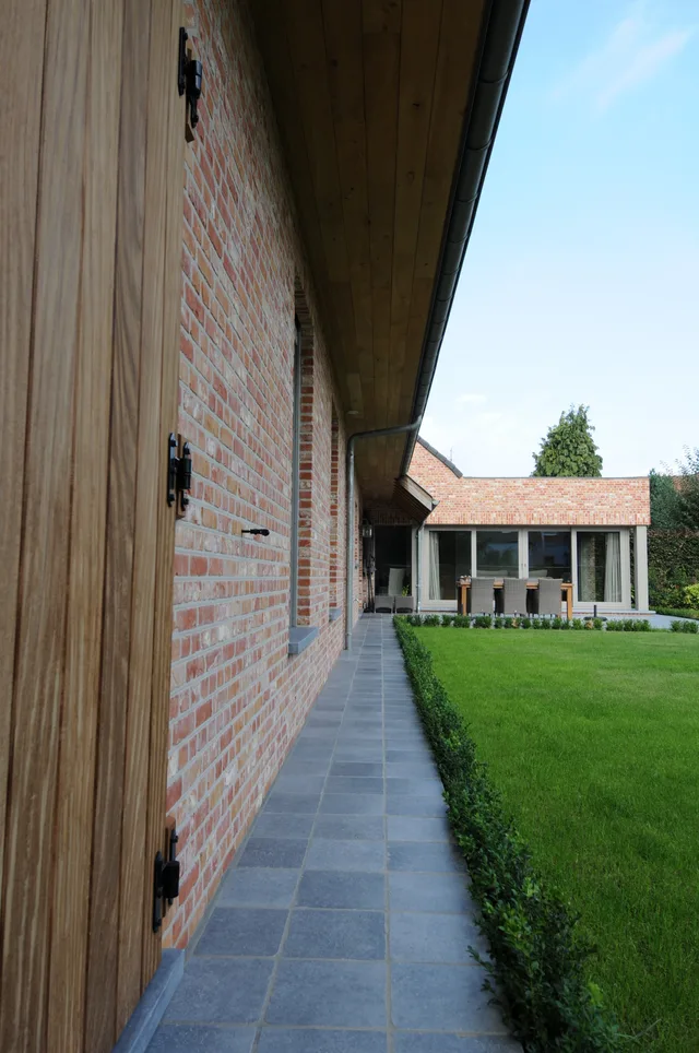 Zijgang met houten deur en bakstenen muur tussen woning en tuin met gazon en haag