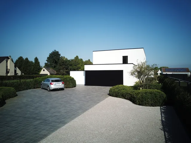 Moderne woning met witte gevelbekleding en zwarte garagedeur, omgeven door aangelegde tuin met groene heesters