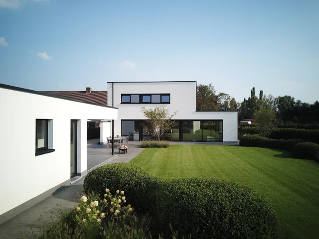 Moderne woning met witte gevel en grote glazen raampartijen, gezien vanaf de tuin met grasveld en haag