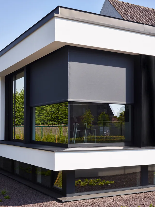 Moderne voorgevel met wit gepleisterde bovenbouw, zwarte aluminium raamprofielen en grijze zonneschermen
