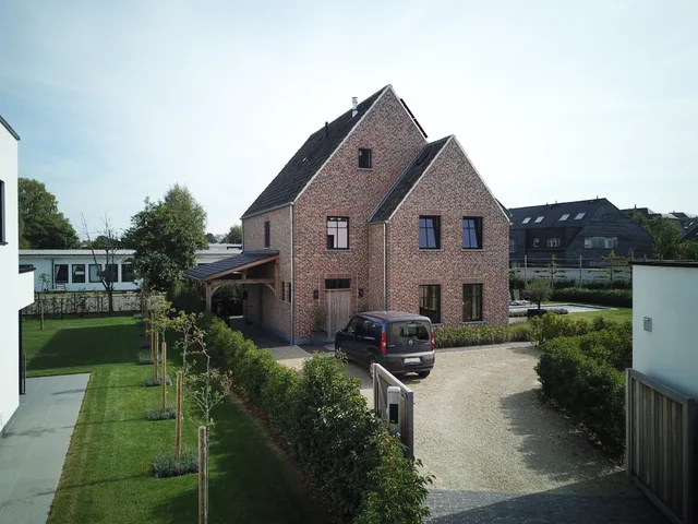Voorgevel van een rode bakstenen woning met dubbele dakkapel, grote ramen en een houten terrasoverkapping