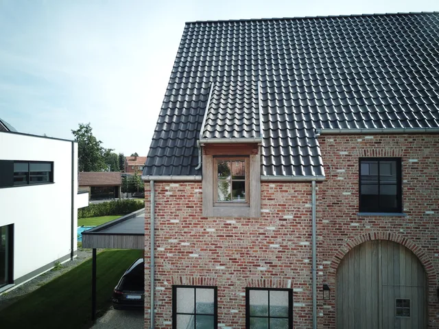 Nieuwbouw woning met dakkapel en antracietkleurig pannendak, naast gerenoveerde bakstenen boerderij