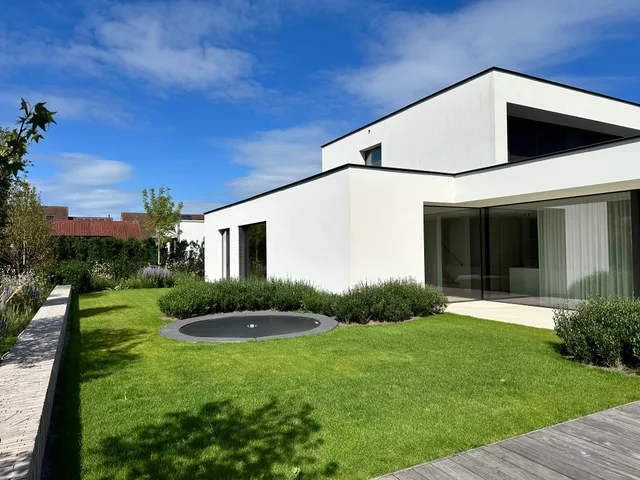 Moderne woning met witte gevel en grote raampartijen, gezien vanuit de tuin met gazon en trampoline