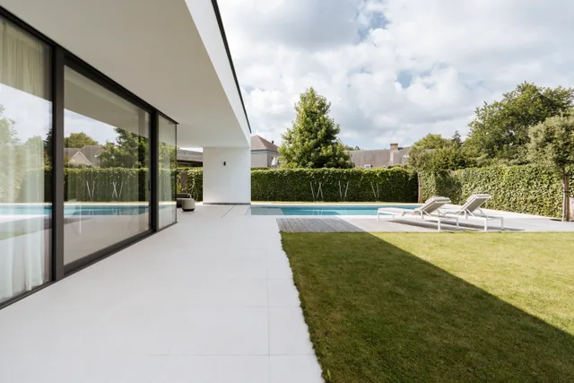 Moderne woning met grote glaspartijen en zwembad, uitkijkend op aangelegd gazon en groene haag