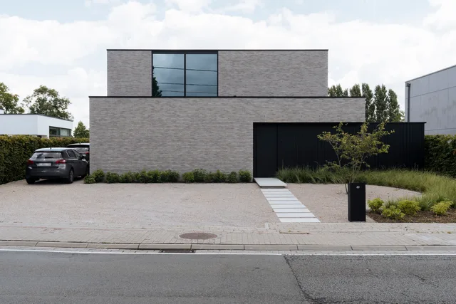 Moderne woning met grijze baksteengevels, grote ramen, zwarte garagedeur en geplaveide inrit met auto