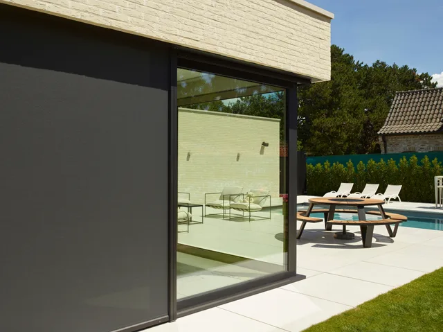 Moderne woning met grote glazen schuifdeuren en terrasoverkapping, uitkijkend op zwembad en tuinmeubelen