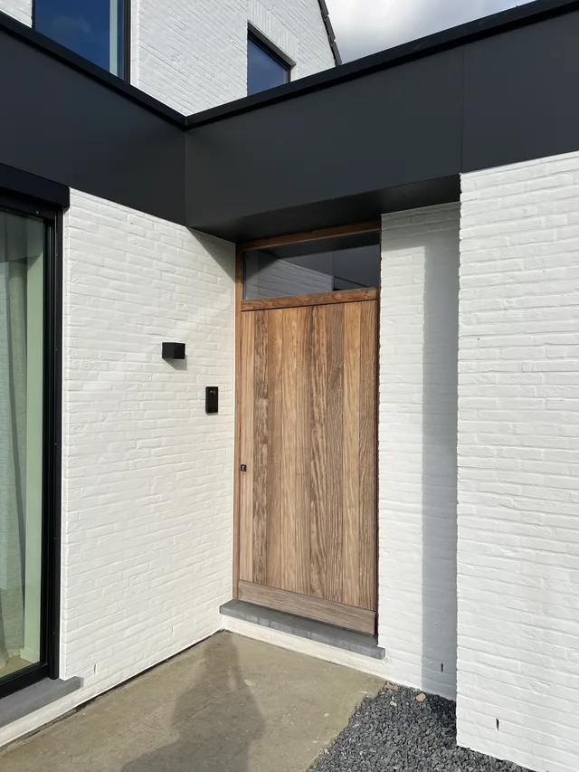 Voorgevel met witte bakstenen afwerking, zwarte overkapping en houten voordeur met glaspartij