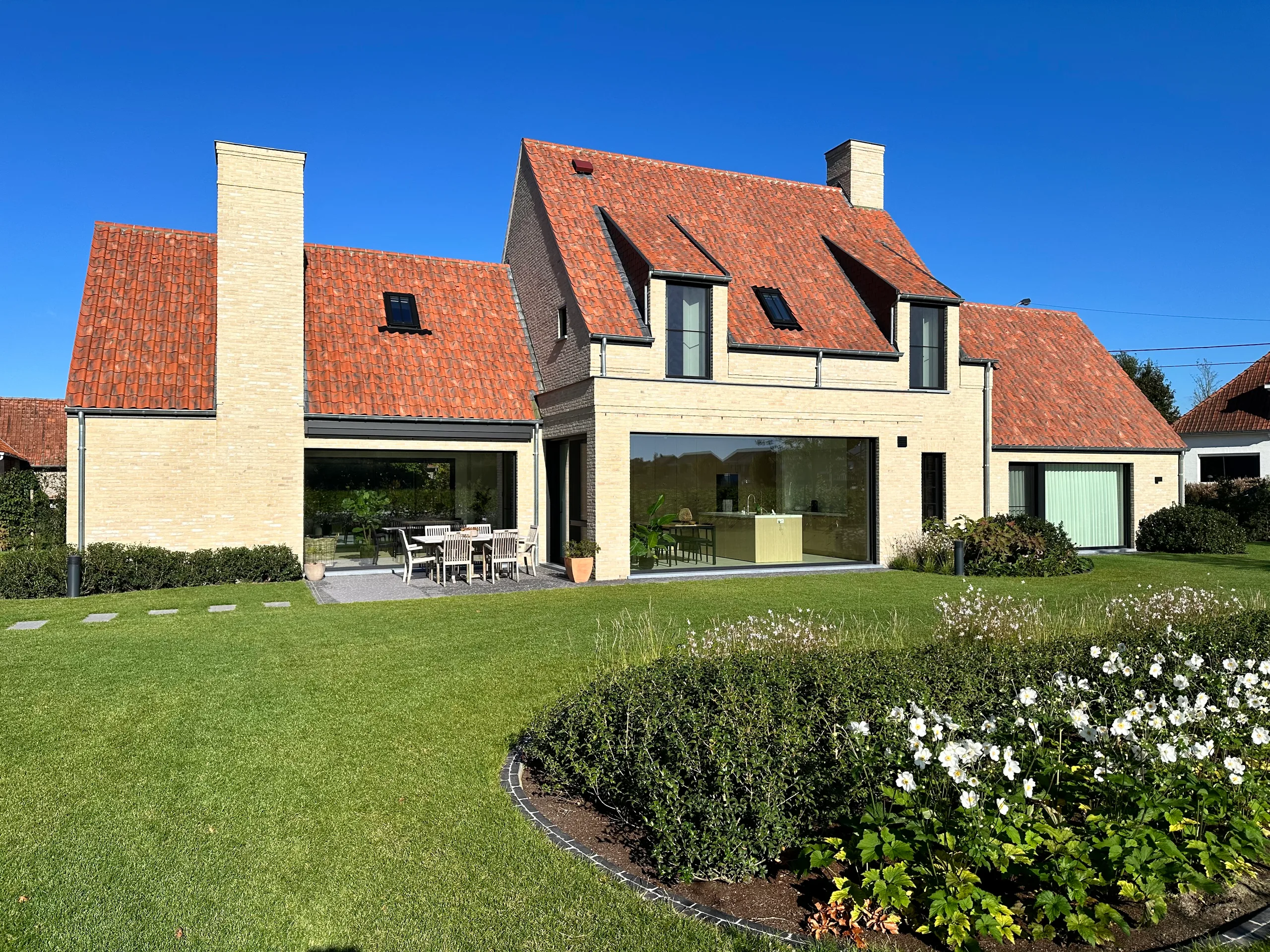 Woning met witte crepi en rode dak, gezien vanaf de tuin met grote glazen puien naar het terras