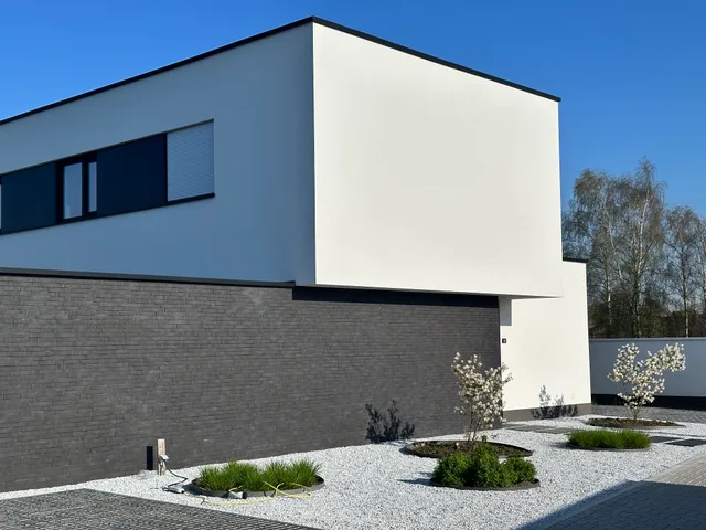 Moderne woning met witte gevel en grijze baksteensokkel, grote raampartijen en strak aangelegde voortuin