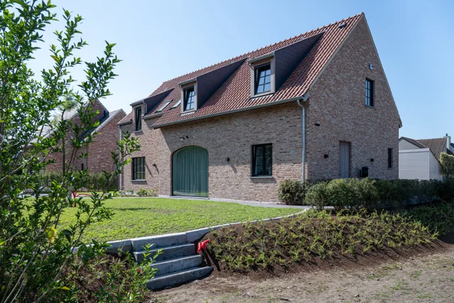 Nieuwbouwwoning met bakstenen gevel, rode dakpannen en dakkapels, omgeven door voortuin met groenvoorziening