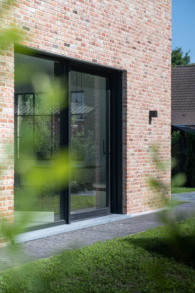 Bakstenen voorgevel met grote zwarte schuifpui die uitgeeft op een terras