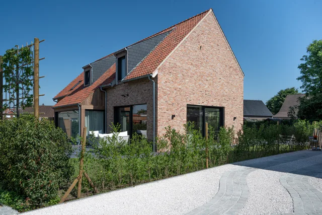 Nieuwbouwwoning met rode bakstenen gevel, dakkapel en grote raampartijen, omgeven door jonge hagen en voortuin