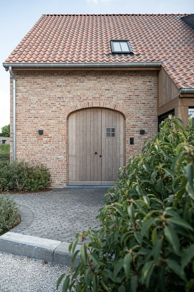 Gevel van een woning met rode bakstenen, oranje dakpannen en houten garagedeuren met gebogen bovenlicht