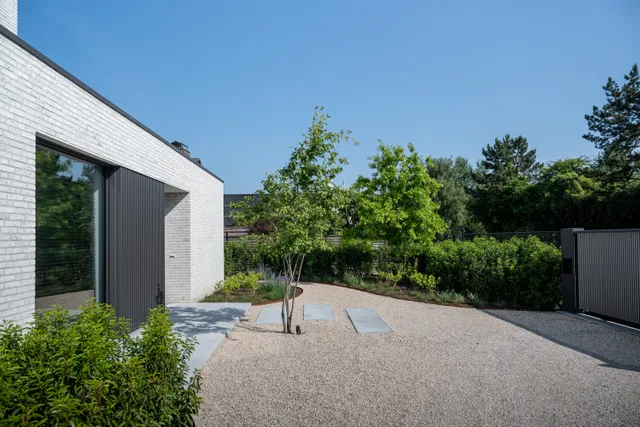 Modern huis met witte gevelbekleding en grote ramen, grijze granitische tuin met groene beplanting en hoge haag