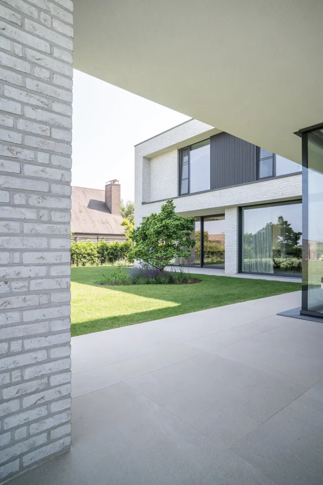 Modern huis met witte bakstenen gevel, grote ramen en terras dat uitgeeft op een tuin met gazon en groene struiken