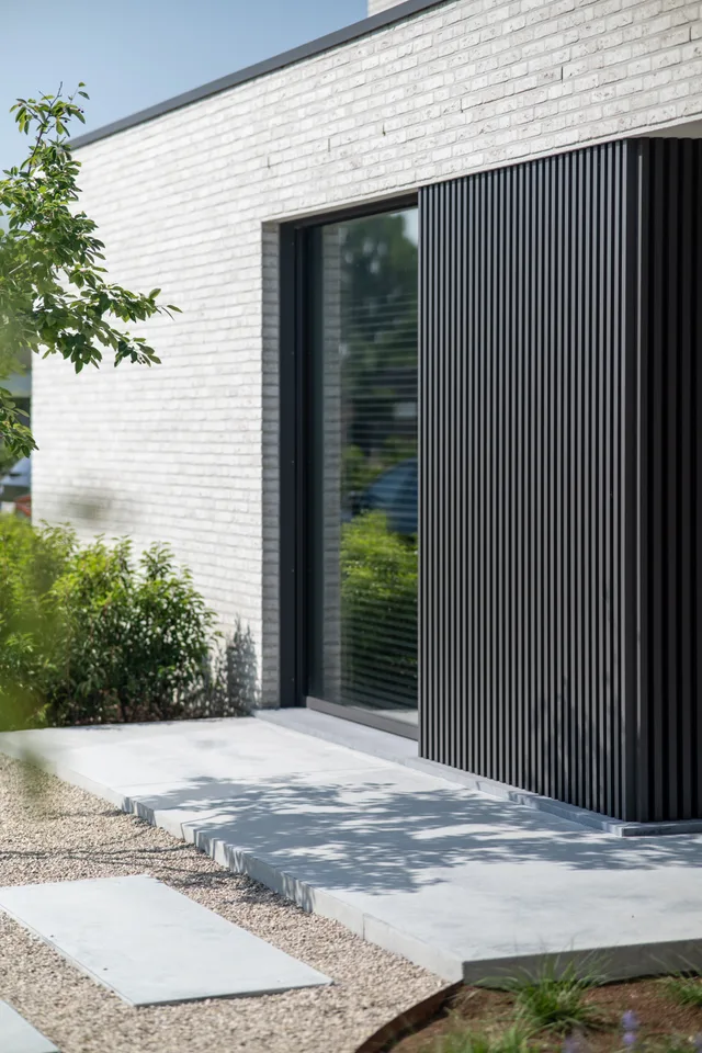 Moderne woning met witte bakstenen gevel, groot raam met zwarte lamellen en verharde ingang met grind