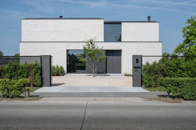 Moderne woning met witgeschilderde gevel, grote raampartijen en een open voortuin met poort en jonge boom