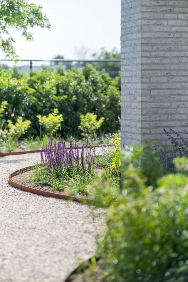 Moderne tuinaanleg met paarse bloemen en stalen borders tegen een bakstenen muur, uitzicht op het landschap