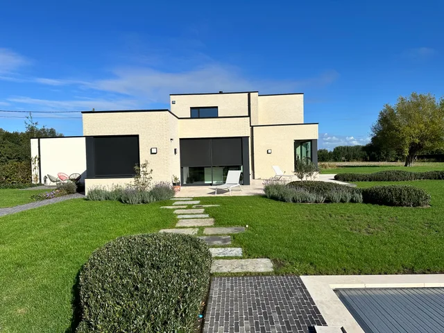 Moderne woning met crème baksteen gevel, grote raampartijen en zwarte luifels, omgeven door aangelegd groen