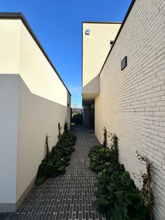 Nauwe toegang tussen twee woningen met gevelsteen en beplanting naar de voordeur