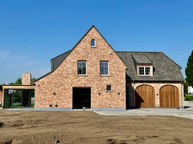 Voorgevel van nieuwbouw boerenwoning in baksteen met dakkapel, houten garagedeuren en moderne aanbouw met grote raampart