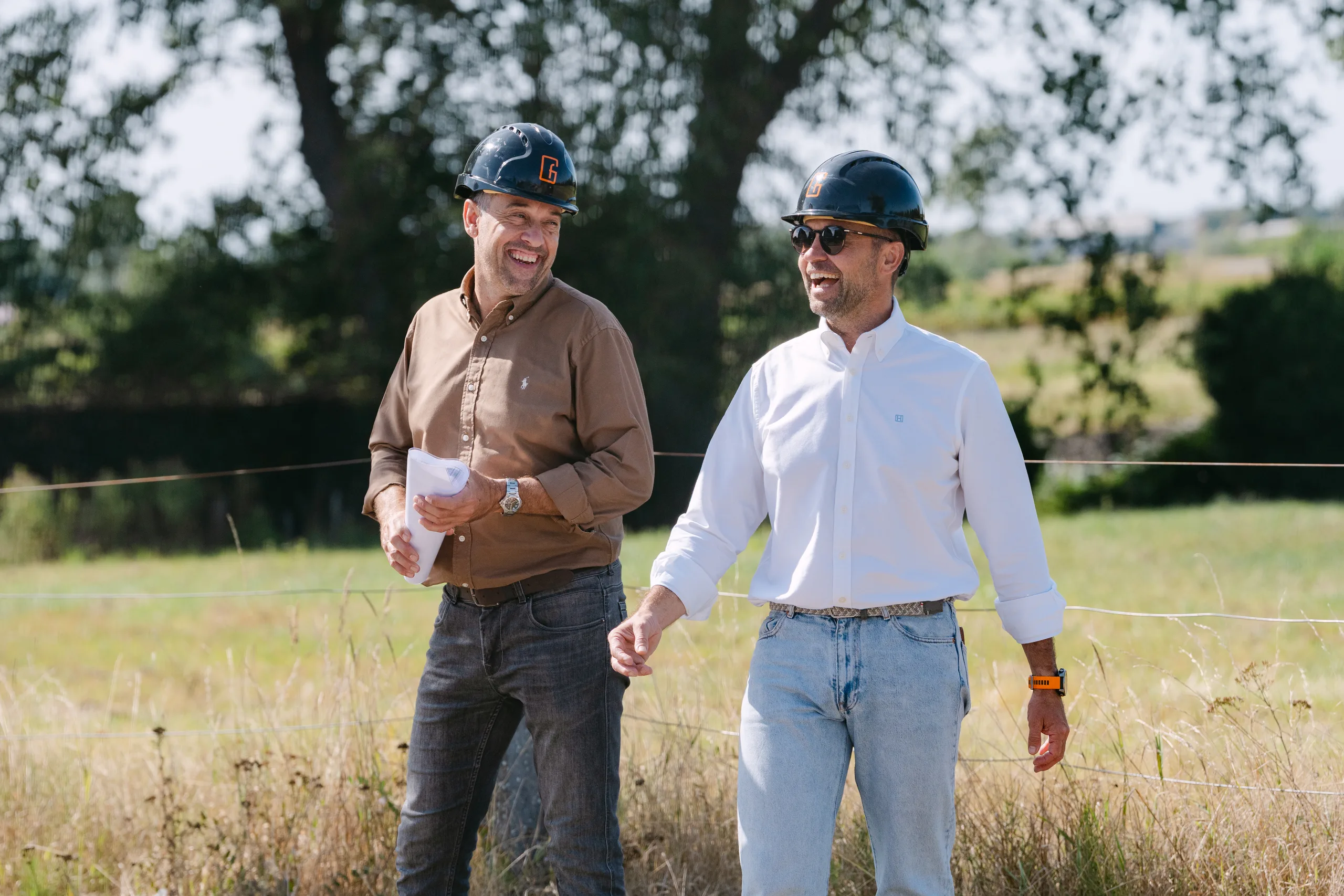 Twee mannen in werkkleding en veiligheidshelmen lopen over een bouwterrein met uitzicht op het landschap