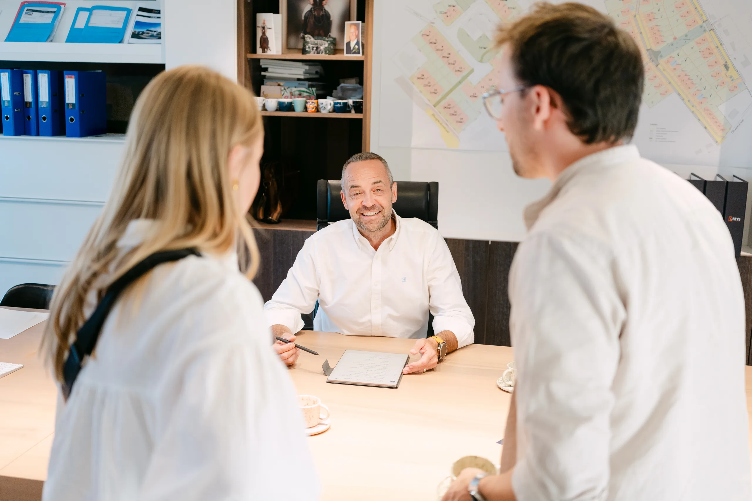 Drie personen in gesprek aan een bureau in een kantoor met kaarten en plannen aan de wand