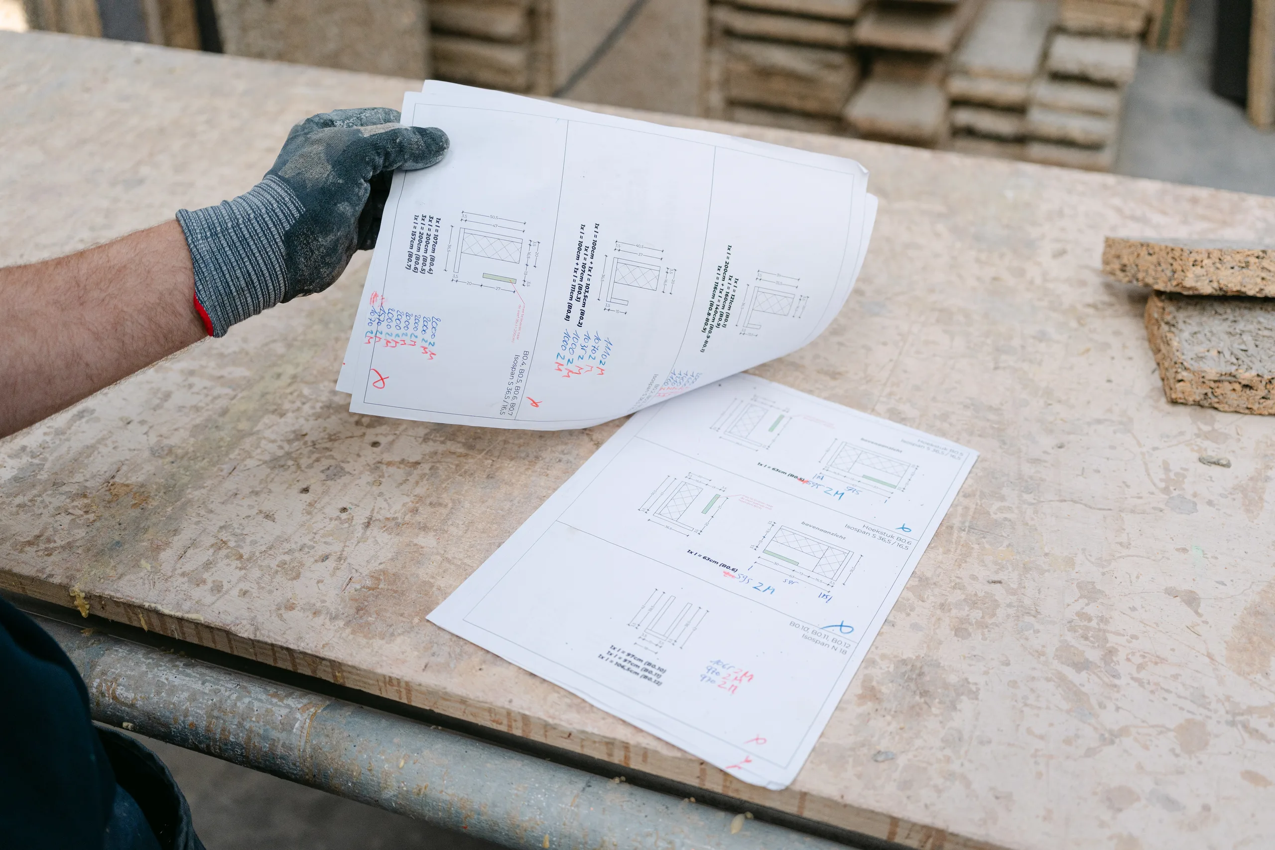 Hand met handschoenen houdt bouwplan vast op werktafel met stapel materiaalstalen op achtergrond
