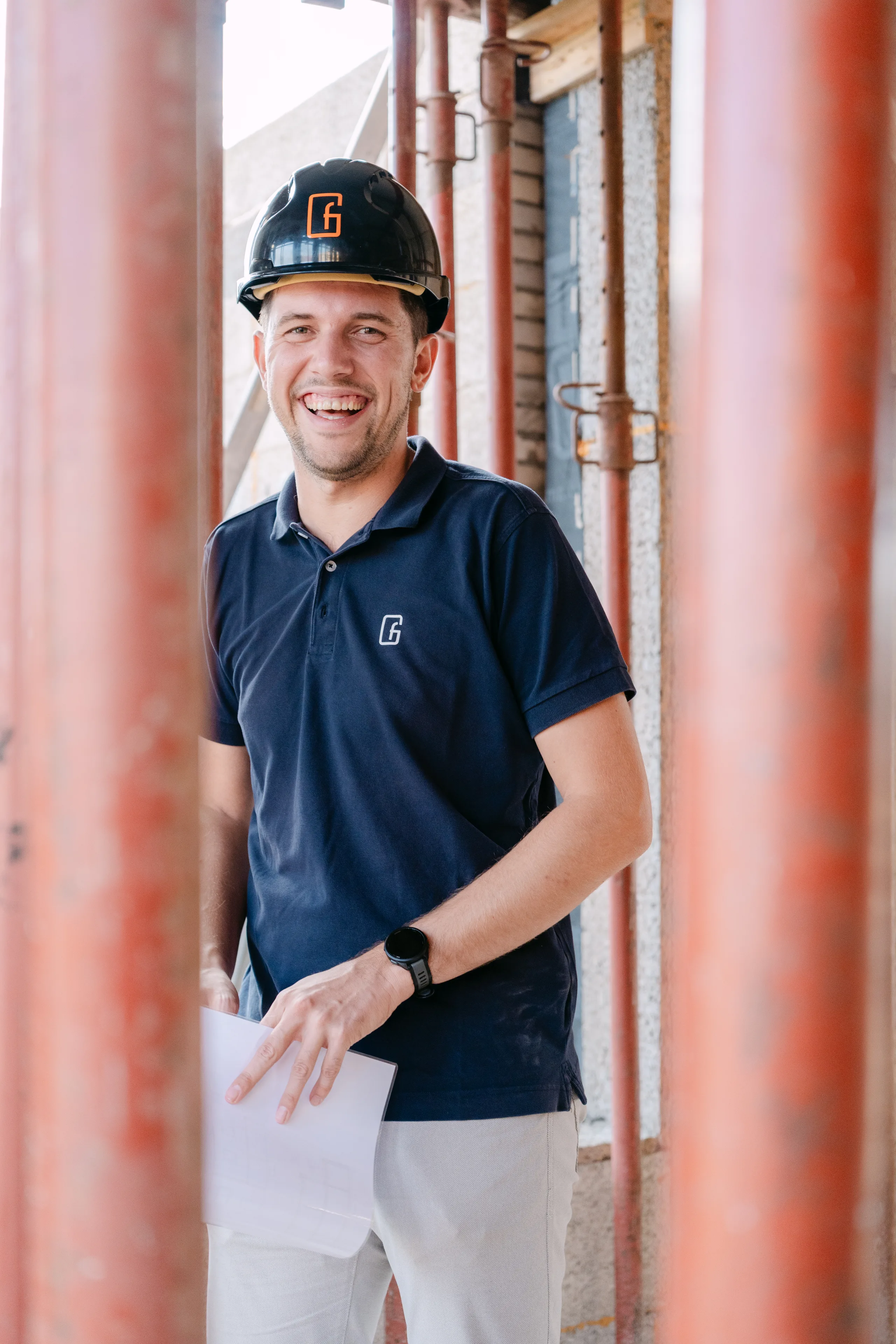 Man met helm en bedrijfskleding op een bouwwerf, staande tussen bakstenen kolommen