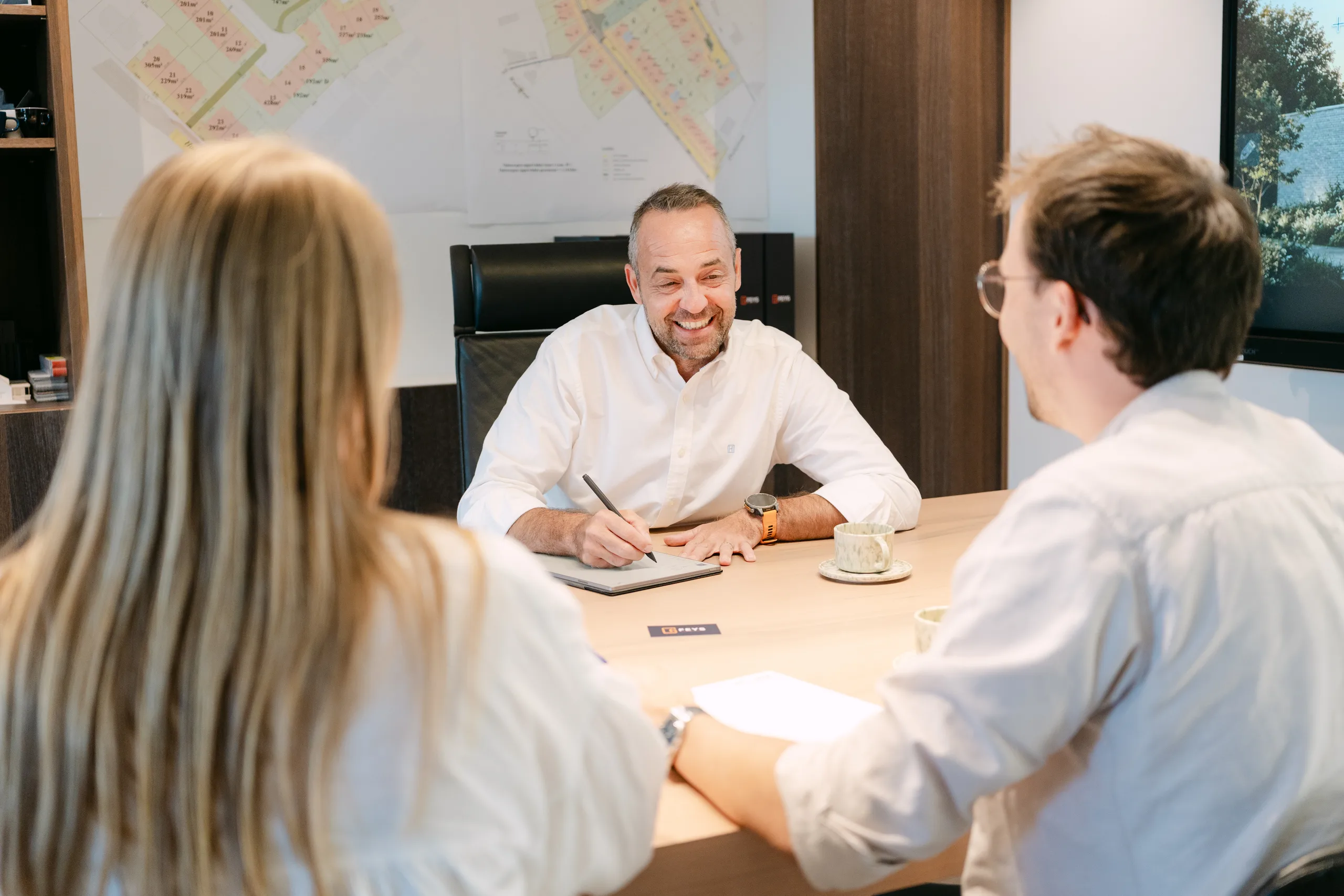 Man en vrouw in gesprek met architect aan bureau met plattegronden aan de muur