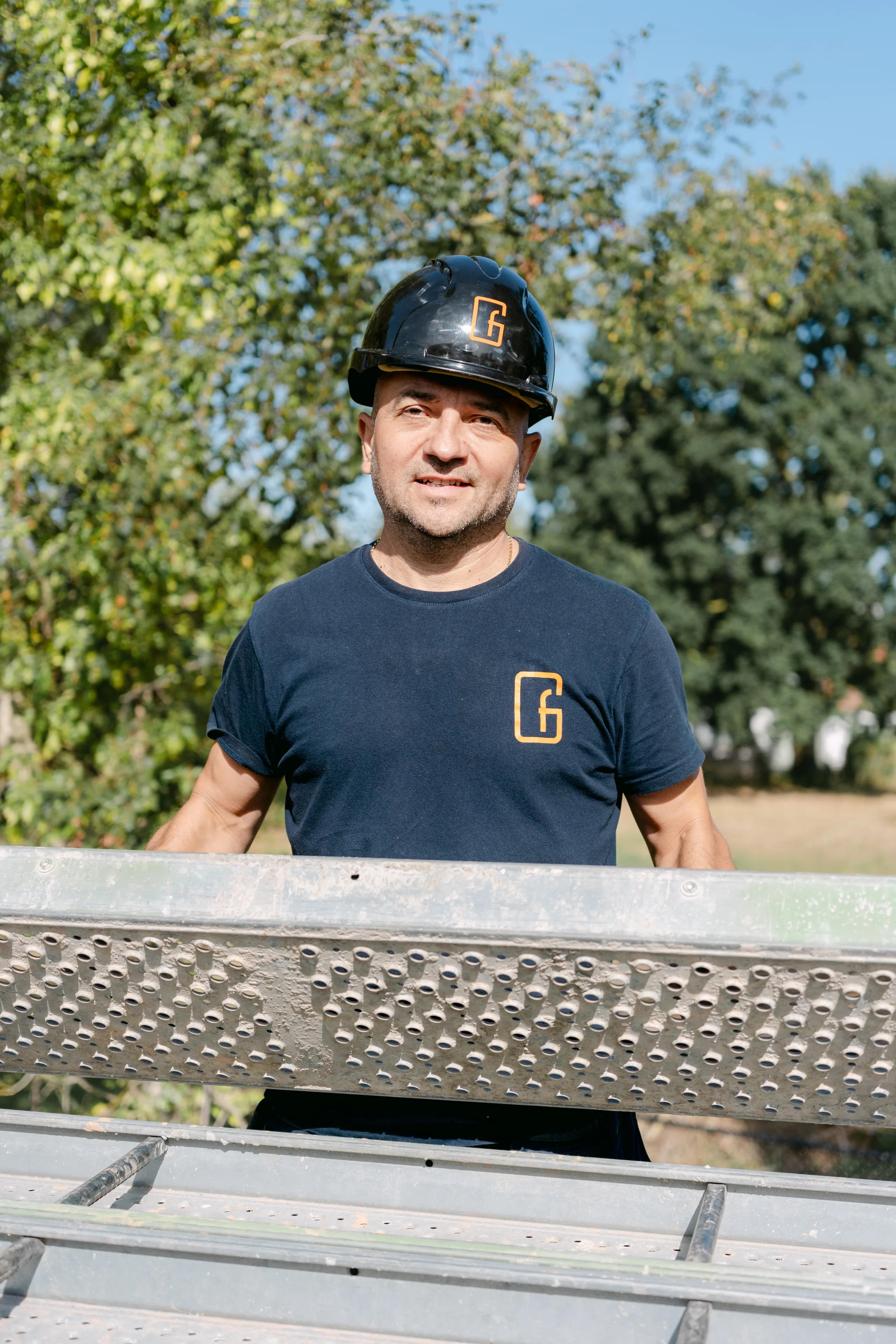 Man met helm en bedrijfslogo op een werf in een groen landschap