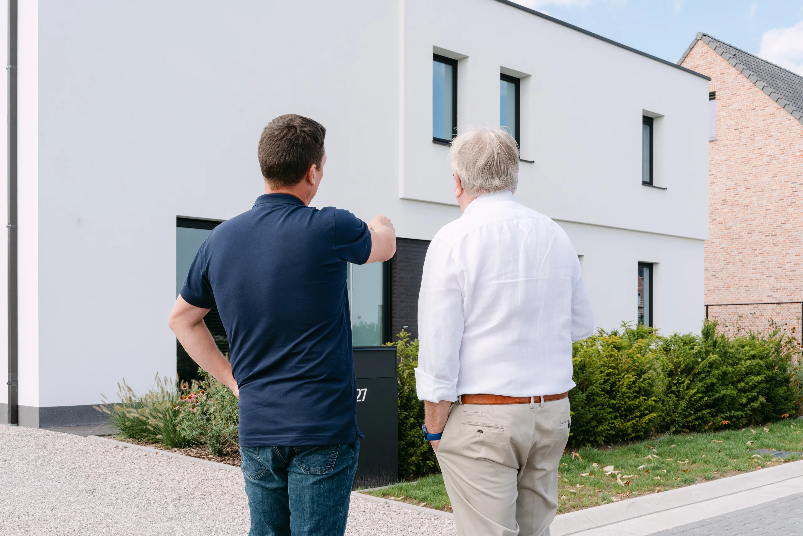 Twee mannen bekijken samen de voorgevel van een moderne woning met witte gevelbekleding en grote ramen