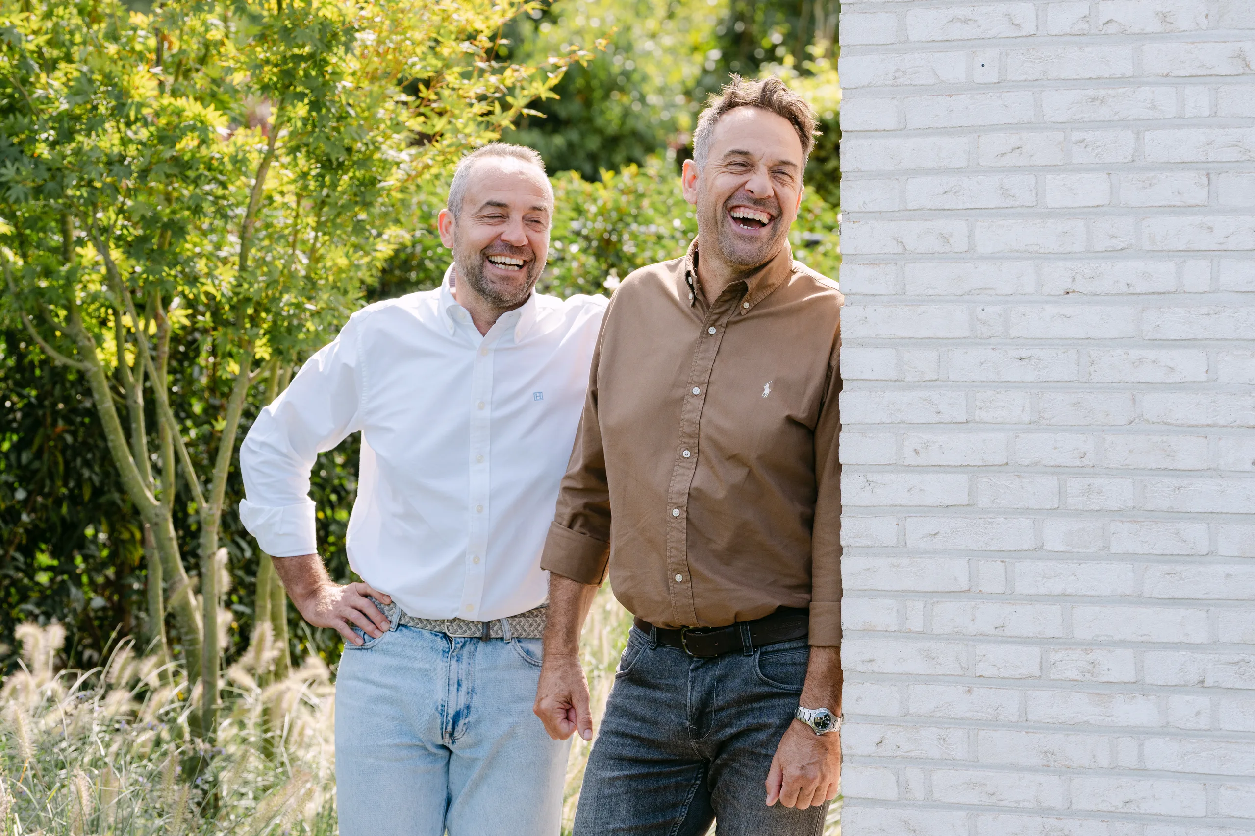 Twee mannen poseren lachend voor een gevel van witgeschilderde baksteen