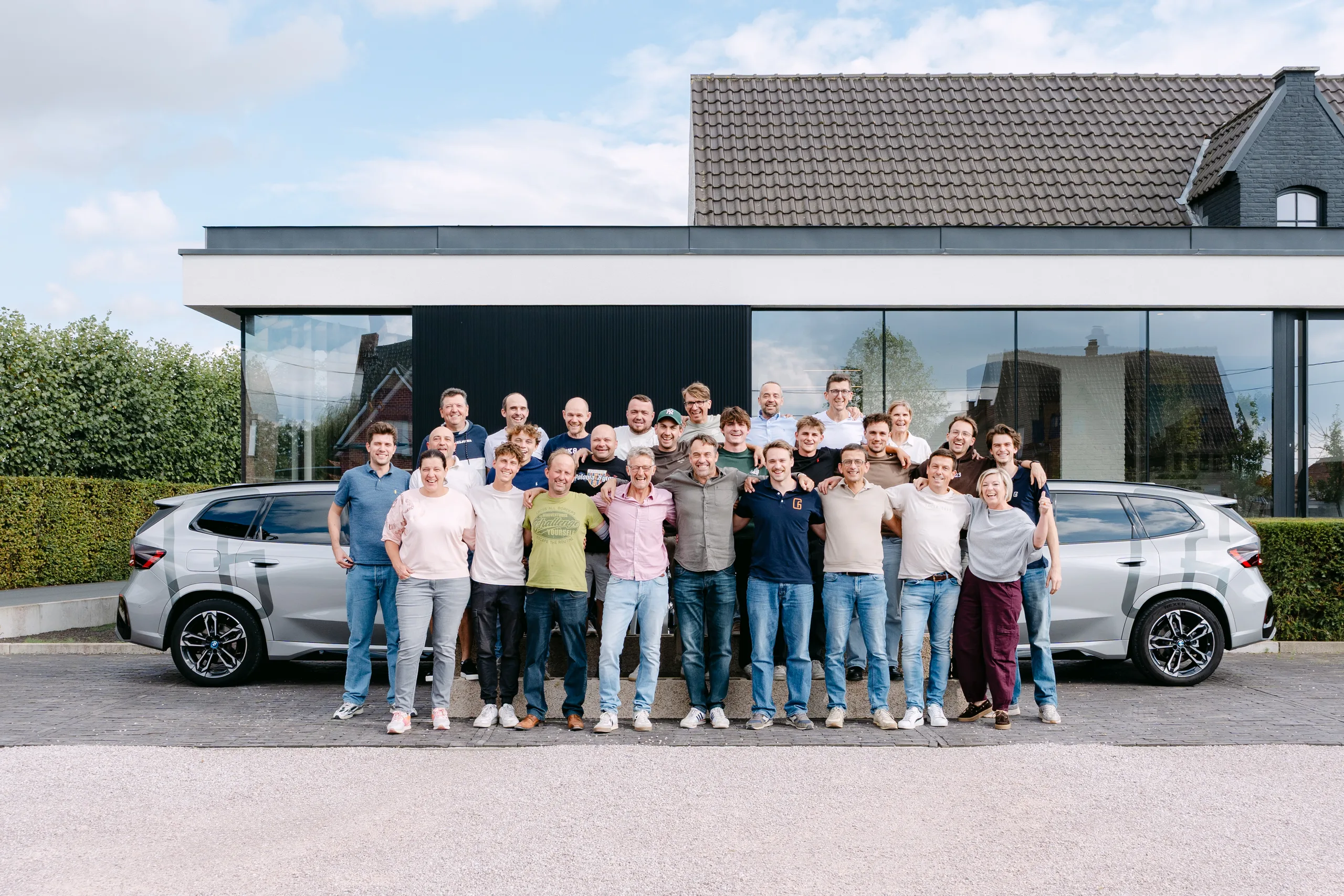 Groepsfoto van meerdere personen voor een moderne villa met uitbouw en twee witte auto's