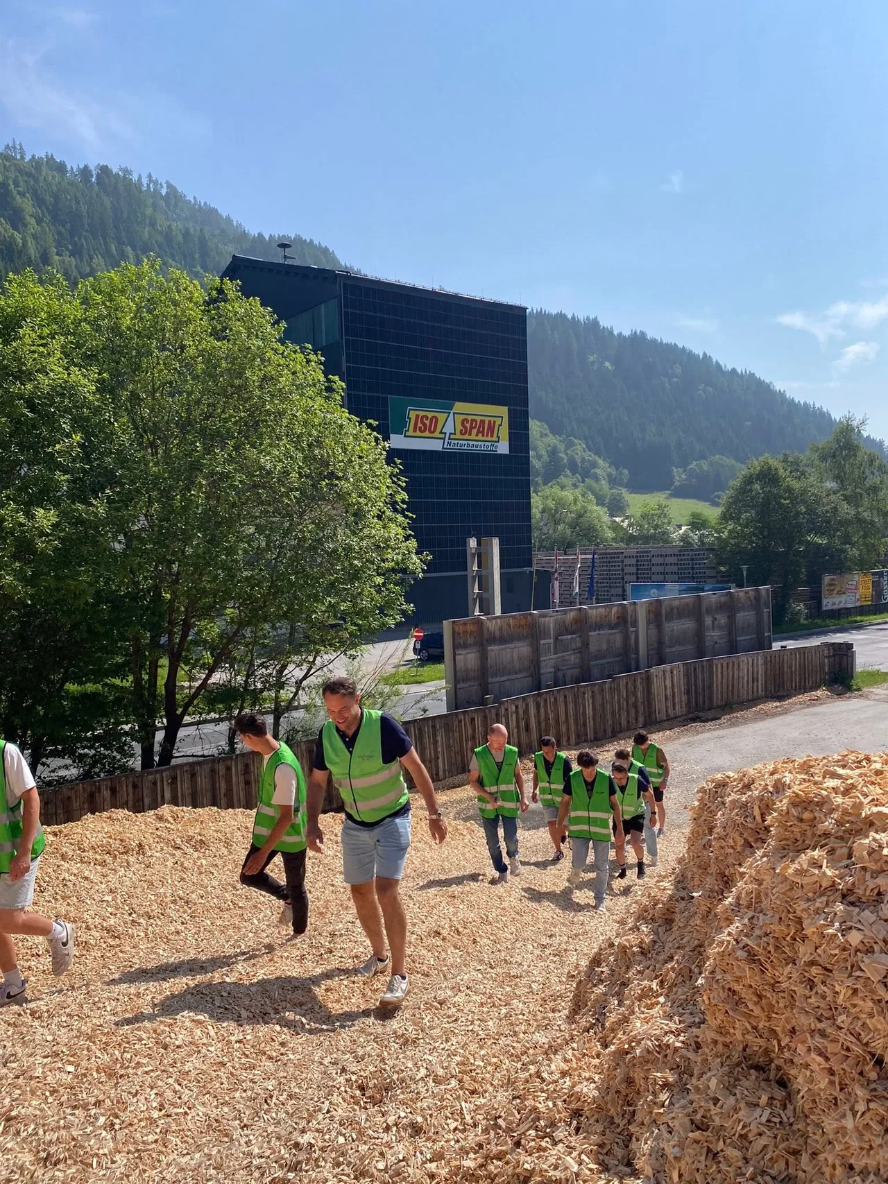 Groep werknemers in hesjes op een bouwterrein met grind en aarden wallen, achter hen een groot zwart gebouw en bosrijke 