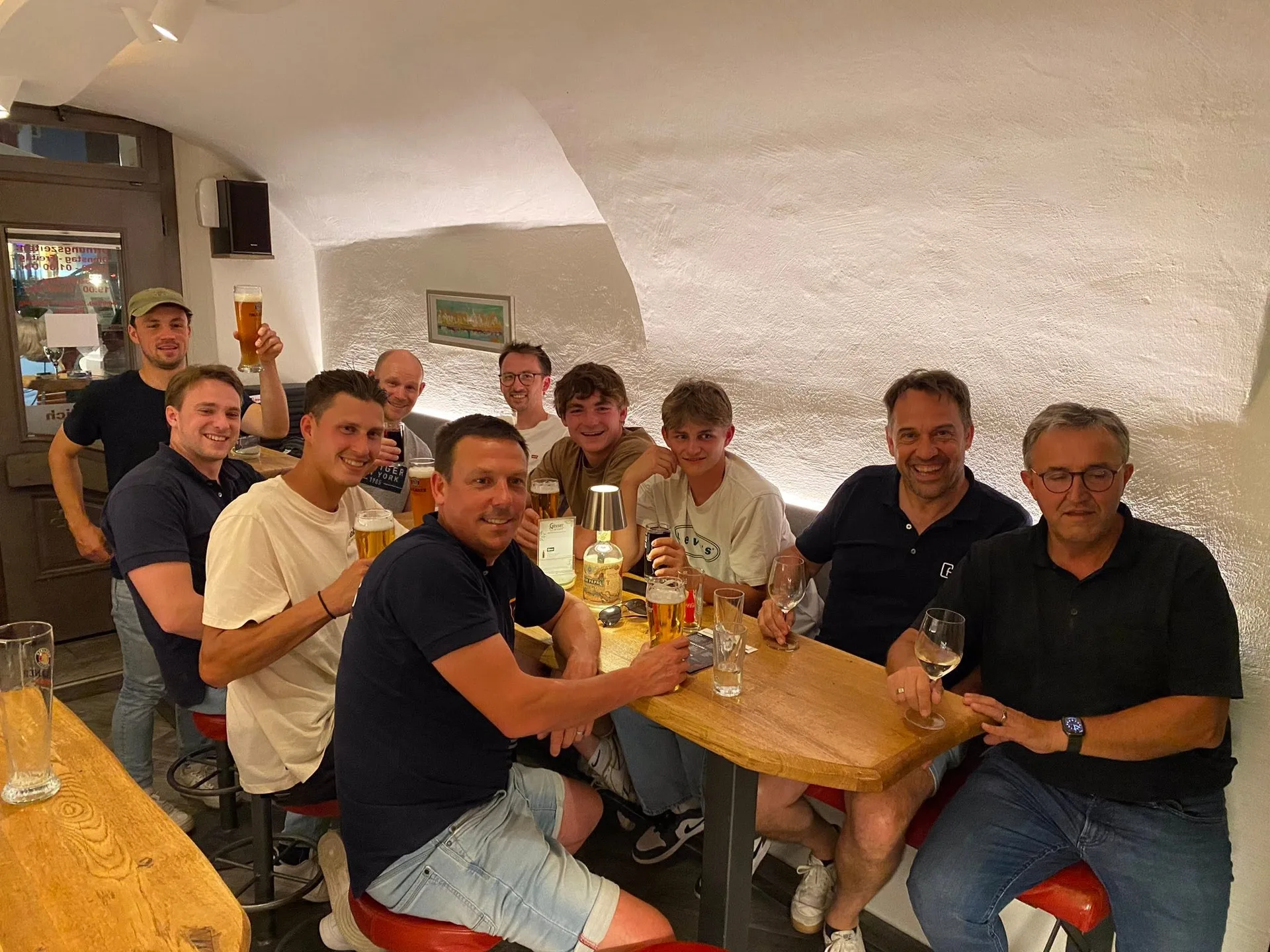 Groep mannen aan houten tafel in gezellige ruimte met witte muren, etend en drinkend