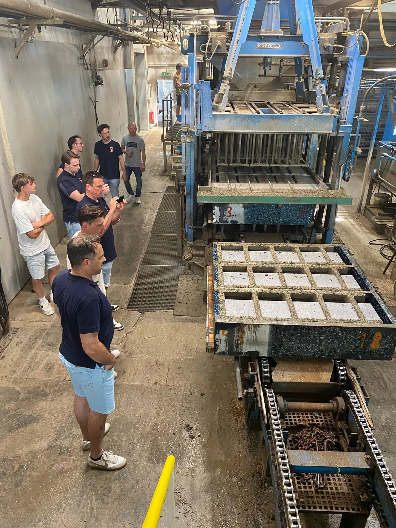 Groep mensen op een bouwwerf kijkt naar een blauwe sorteer- of zaagmachine met betonblokken