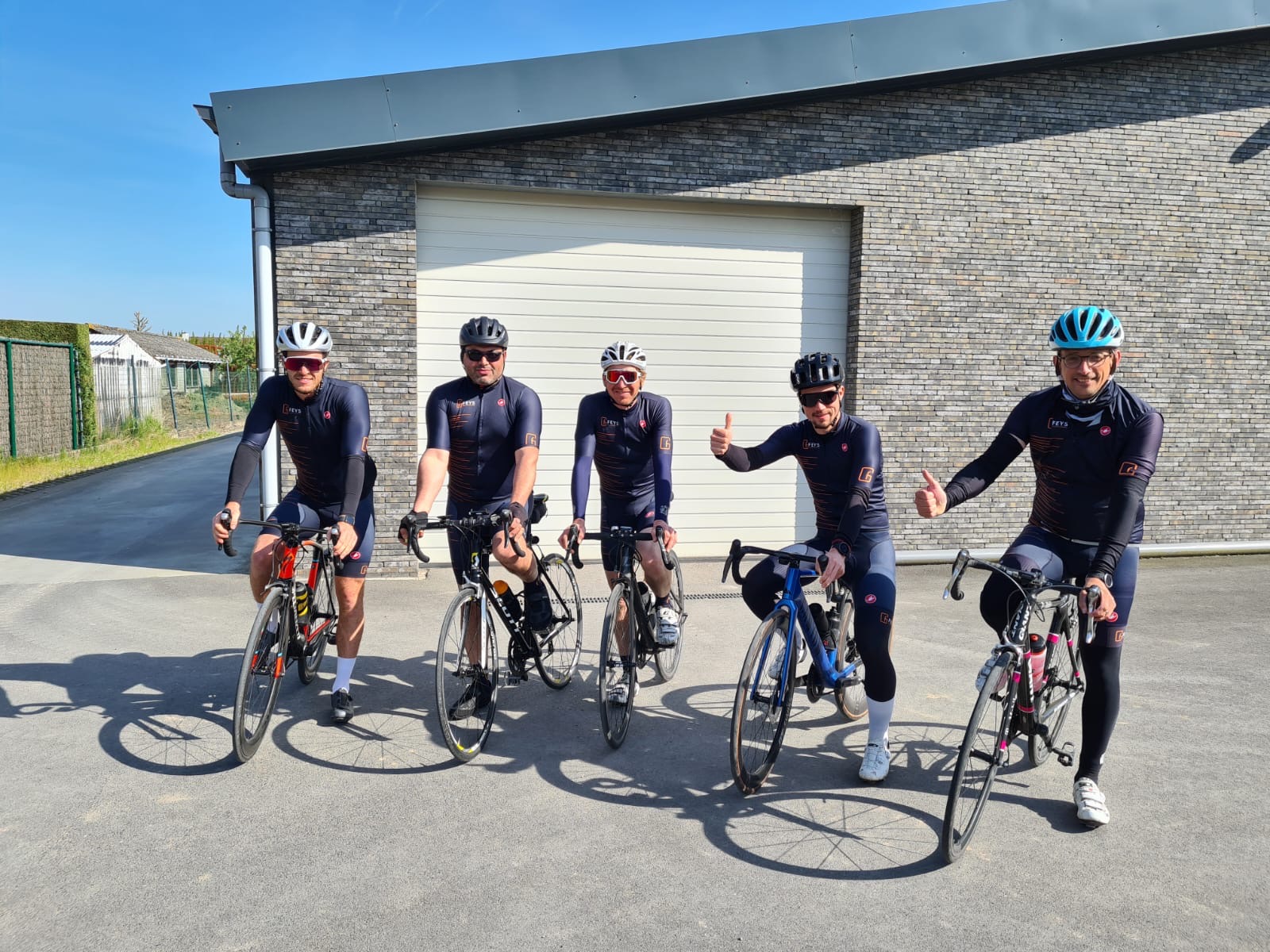 Vijf wielrenners poseren met hun racefietsen voor een moderne garage met bakstenen gevel