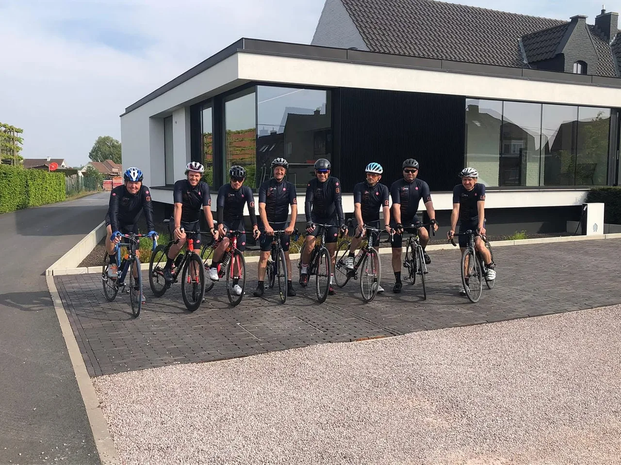 Groep fietsers in teamoutfit poseren voor een moderne woning met grote raampartijen