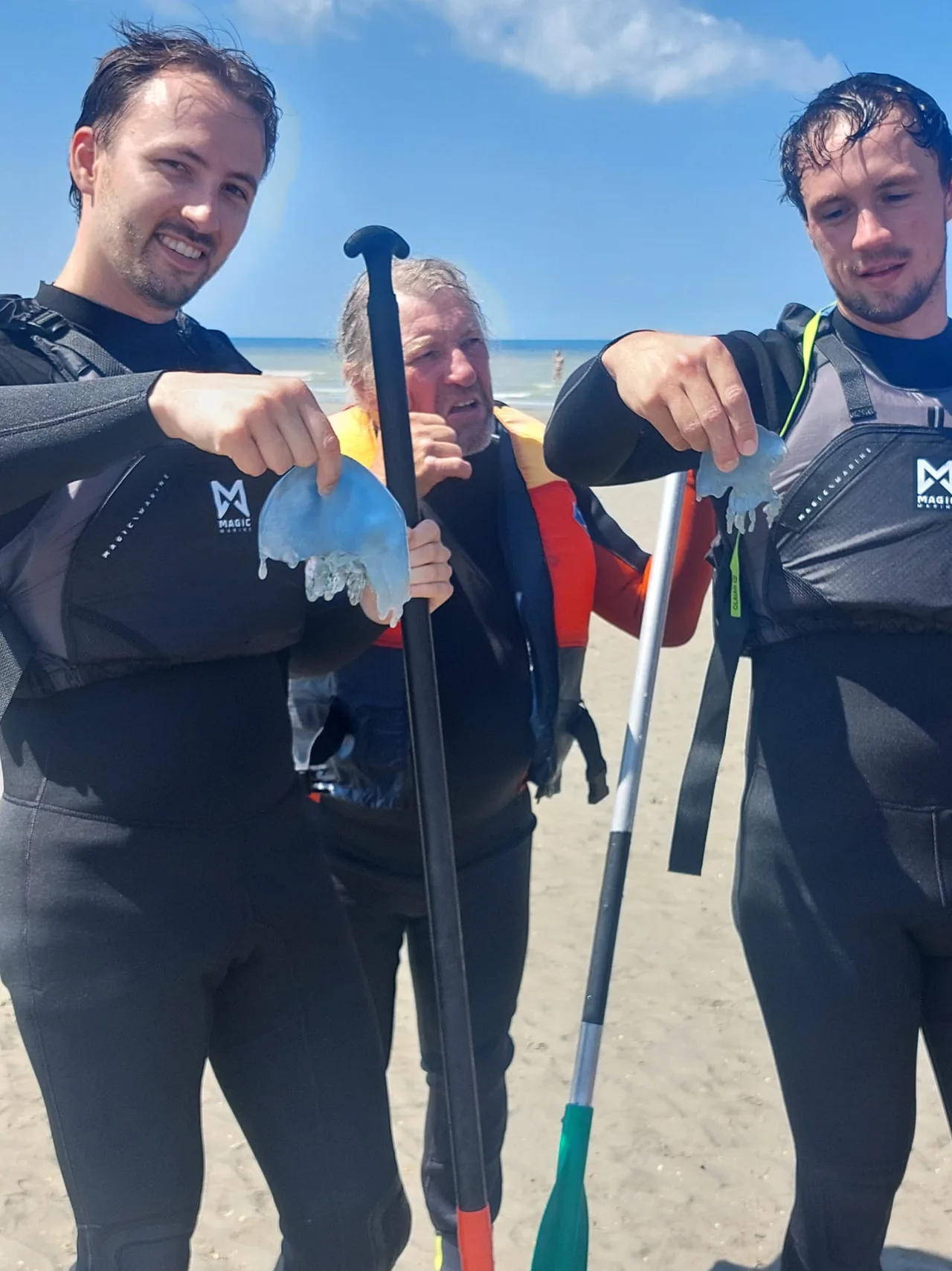 Drie personen in wetsuits op het strand met peddels en watersporten uitrusting