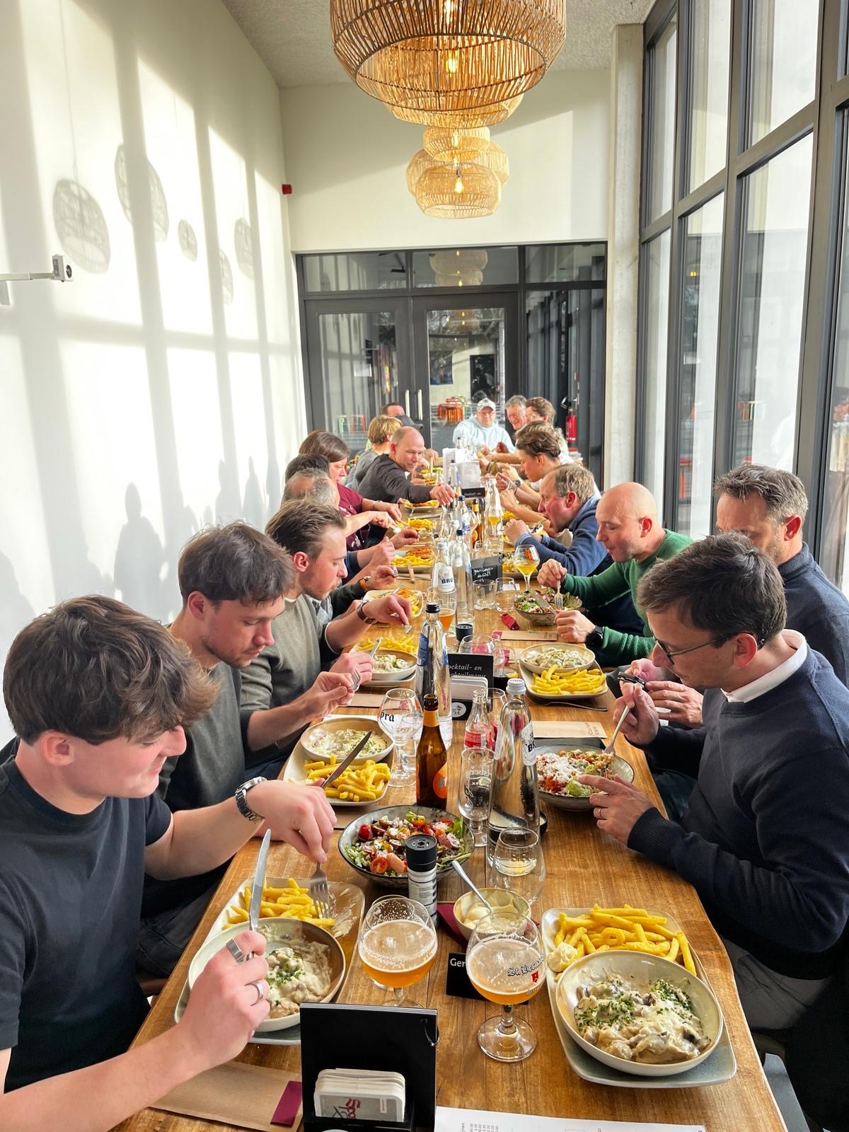 Groep mensen eet samen aan een lange houten tafel in een lichte ruimte met grote ramen en hangende lampen