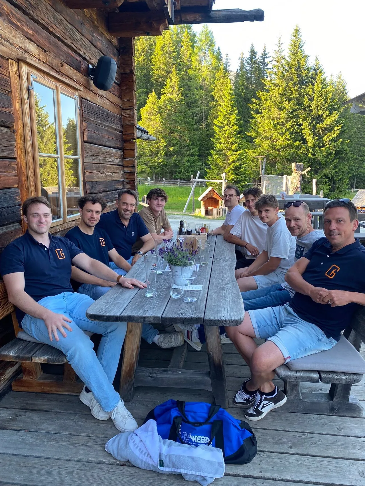 Groep mensen zit gezellig aan een houten tafel op een houten terras tegen een houten chalet, omgeven door bos