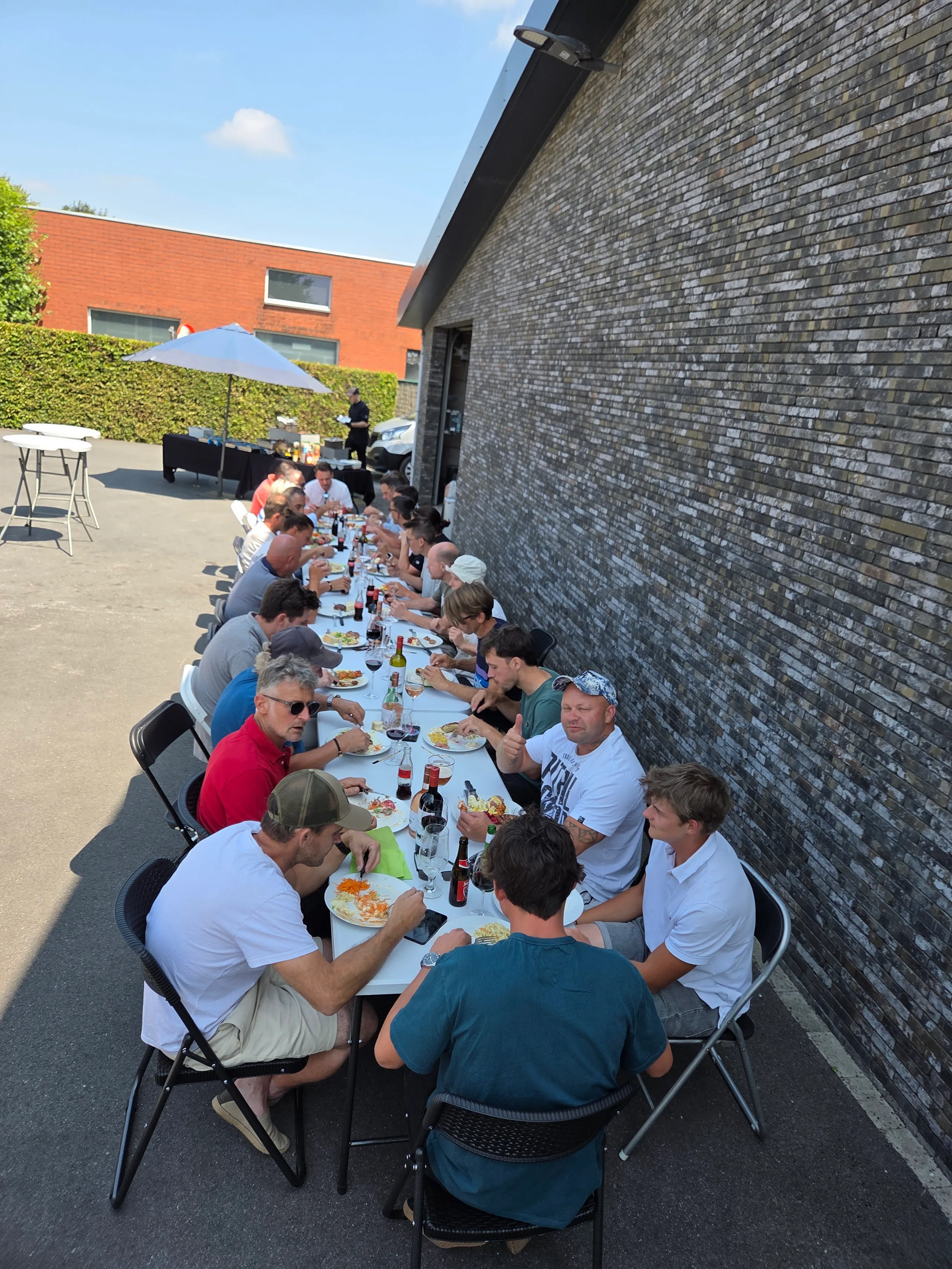 Groep mensen eet aan lange tafel op binnenplaats tegen grijze bakstenen gevel