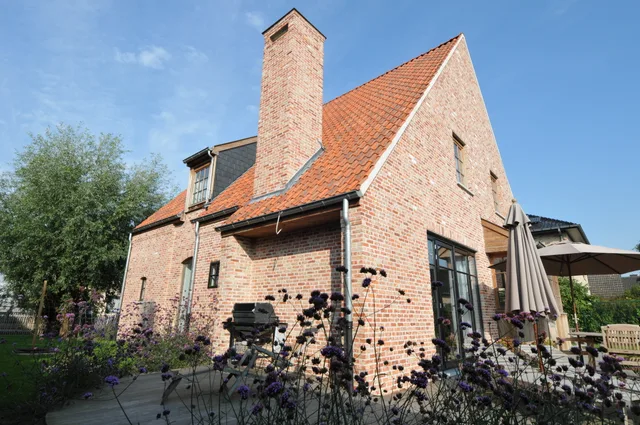 Voorgevel van een bakstenen woning met oranje dakpannen, schoorsteen en tuinplanten op het terras